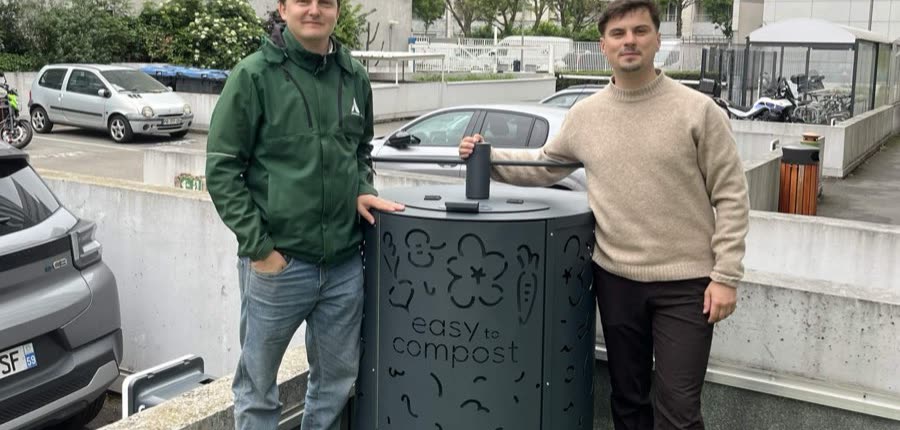 Zwei Mitarbeitende von Easy to Compost beiderseits eines anthrazitgrauen ABC-Komposters mit Logo, auf einem Stellplatz in einem städtischen Hof, zwischen den Fahrzeugen und einem Fahrradraum, ohne jeden Anschluss