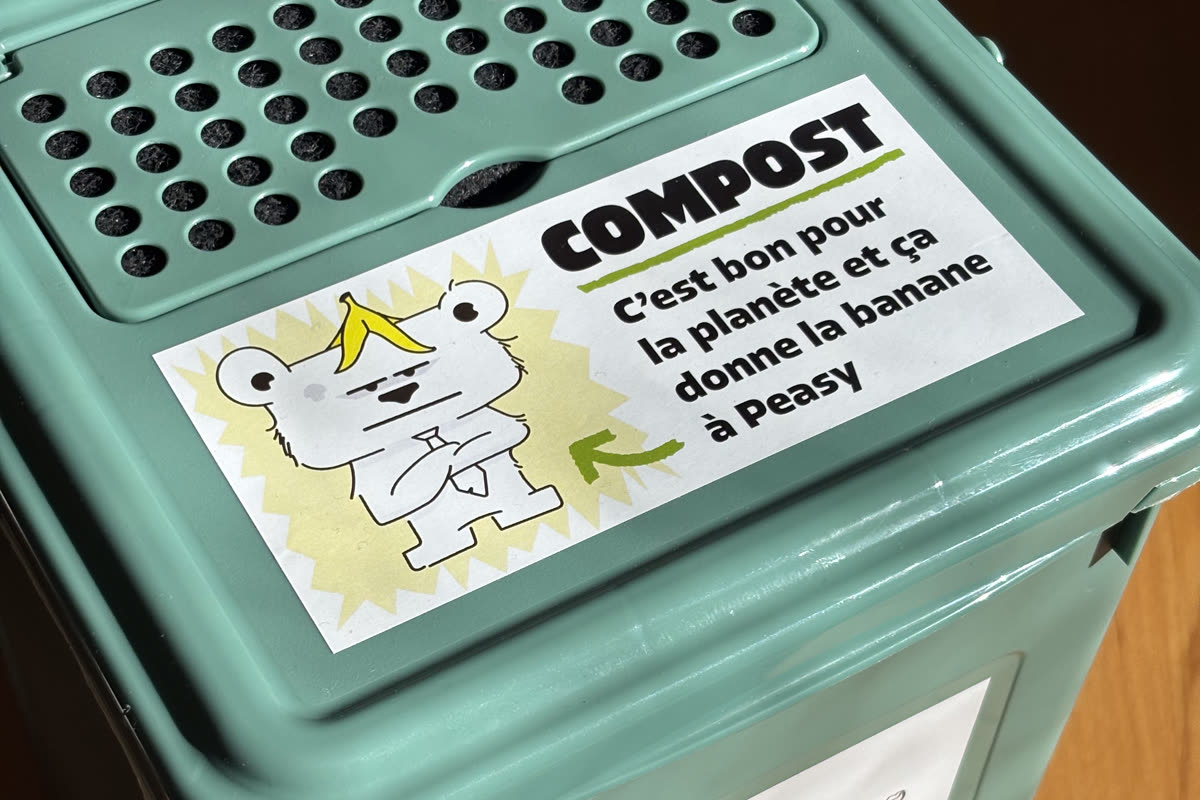 Deckel eines grünen Vorsortiereimers mit rund sechzig Löchern und dahinterliegendem schwarzem Aktivkohlefilter, dazu ein Aufkleber zur Sensibilisierung für die Bioabfalltrennung.